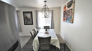 Un comedor con una mesa dispuesta para cuatro, una araña, cortinas estampadas y un cuadro en la pared.