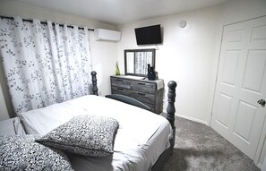 Un dormitorio con cama, cómoda, televisor montado en la pared y una puerta.