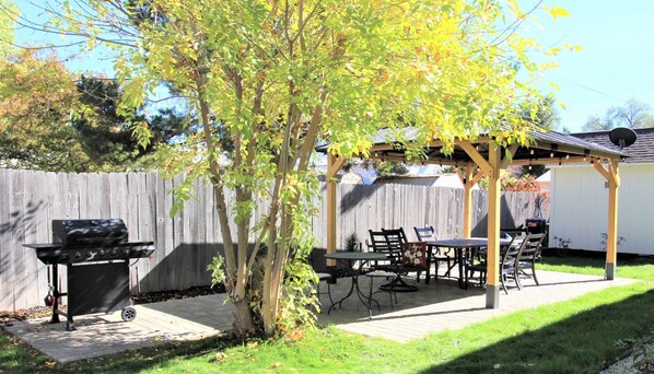 A backyard with a wooden pergola, a barbecue grill, and a dining table with chairs.