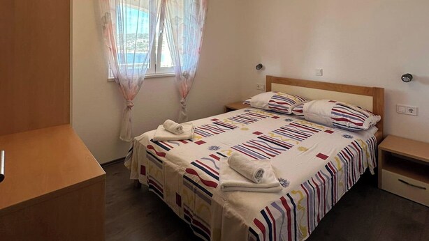 Ein Schlafzimmer mit einem Bett, einem Nachttisch, einem Holzschrank, einem Fenster mit leichtem Vorhang und einer Deckenleuchte.