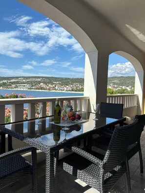 A glass-top table with wicker chairs, a bottle, and fruit on a balcony overlooking a coastal town. - 2-bedroom apartment in peaceful Sevid na moru with AC (Sevid na moru)