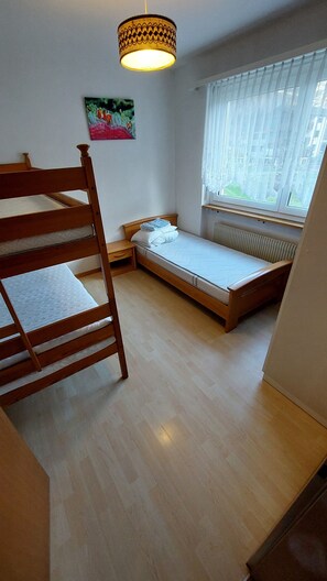 Ein Zimmer mit Etagenbett, einem Einzelbett, einem Heizkörper, einem Fenster mit durchsichtigen Vorhängen und einer Pendelleuchte.