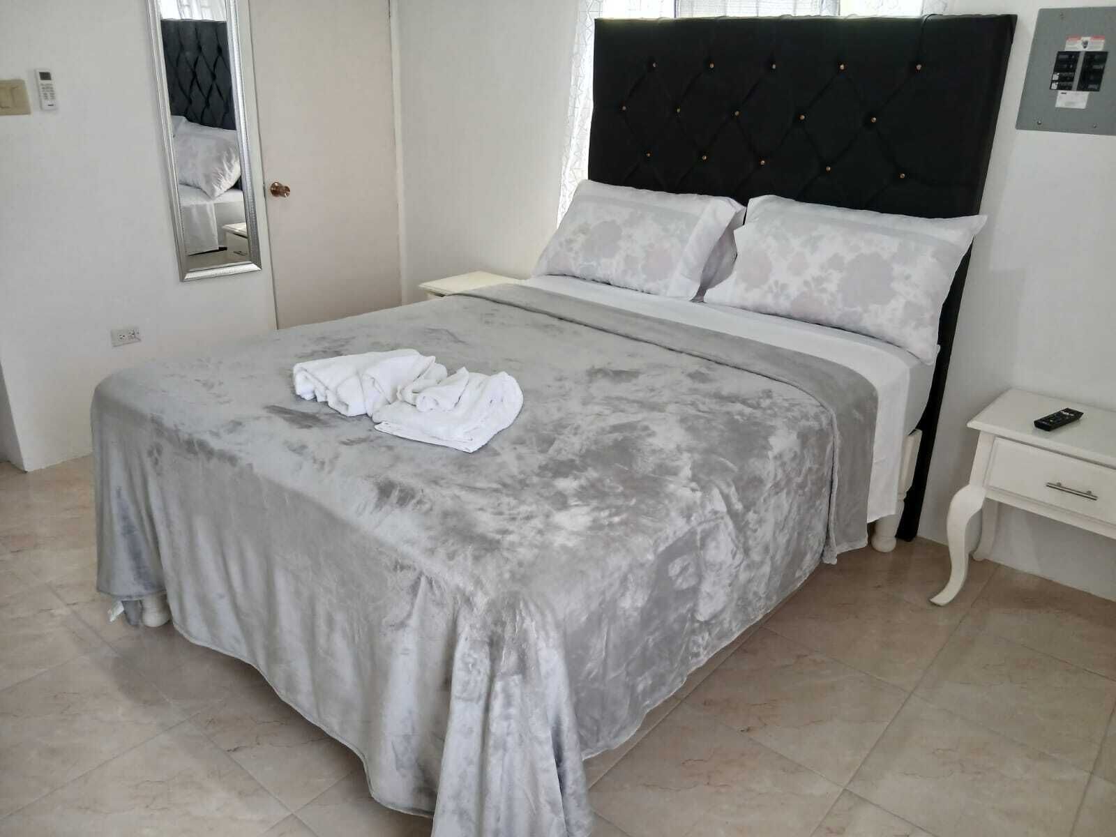 A neatly made bed with a grey bedspread and white pillows, a bedside table with a remote control, and a mirror on the wall.