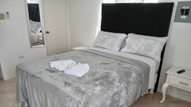 Una cama bien hecha con una colcha gris y almohadas blancas, una mesita de noche con un control remoto y un espejo en la pared.
