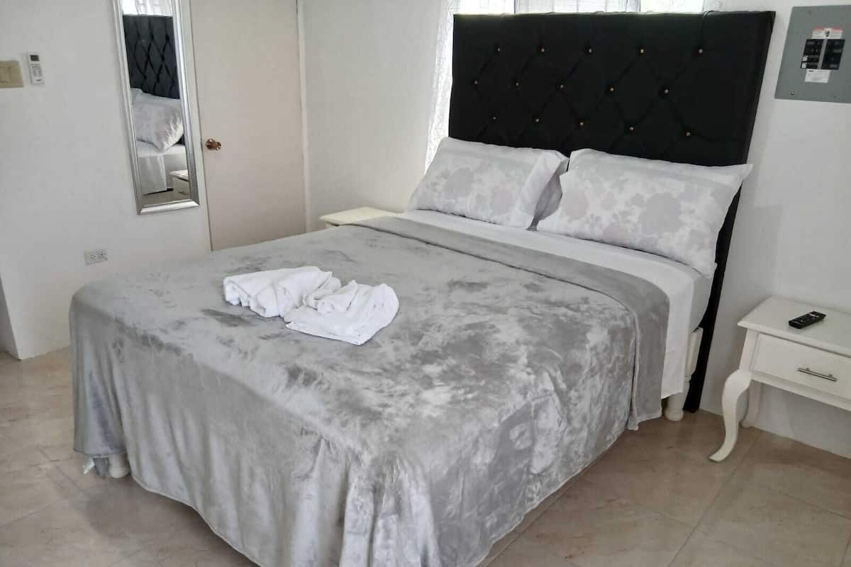 Una cama bien hecha con una colcha gris y almohadas blancas, una mesita de noche con un control remoto y un espejo en la pared.