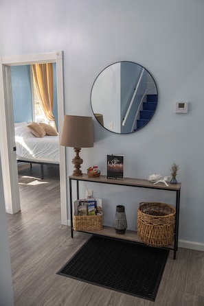 Una habitación de hotel con una cama, una mesita de noche con lámpara, un espejo, un cartel de bienvenida enmarcado, una pequeña cesta, un florero y una cesta de mimbre.