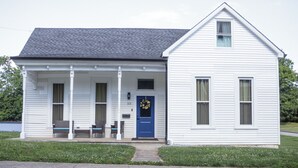 A white single-story house with a blue front door, a porch with wicker furniture, and a small window on the right side. - Close to Mammoth Cave and other wonderful attractions yet still in a quiet area. (Horse Cave)