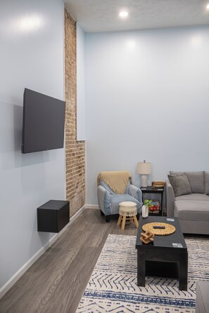 A modern living room with a flat-screen TV mounted on the wall, a grey sofa, a small wooden stool, a coffee table with a woven placemat, and a patterned rug. - Close to Mammoth Cave and other wonderful attractions.  location. (Horse Cave)