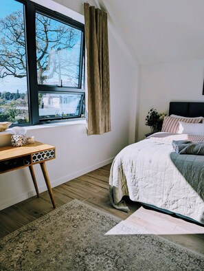 Un dormitorio con cama, escritorio, ventana con cortinas y vistas a árboles y edificios.