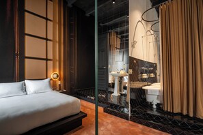 Habitación de hotel moderna con una cama grande, un separador de vidrio, un baño con lavabo e inodoro, y un espejo.