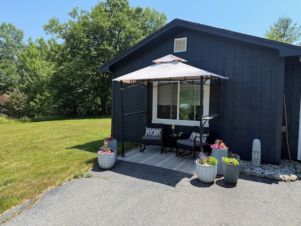 A dark wooden cabin with a covered patio, outdoor seating, and potted plants. - Starry Night Studio, cozy studio in Ellsworth Maine. Pet Friendly! (Ellsworth)