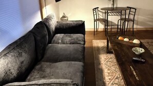 A living room with a sectional sofa, a wooden coffee table, a dining table with chairs, a wall-mounted TV, and a window with blinds.