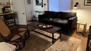 A living room with a dark sofa, a coffee table with pumpkins, a metal shelf unit, and a window with blinds.