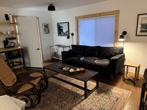 A living room with a dark sofa, a coffee table with pumpkins, a metal shelf unit, and a window with blinds.