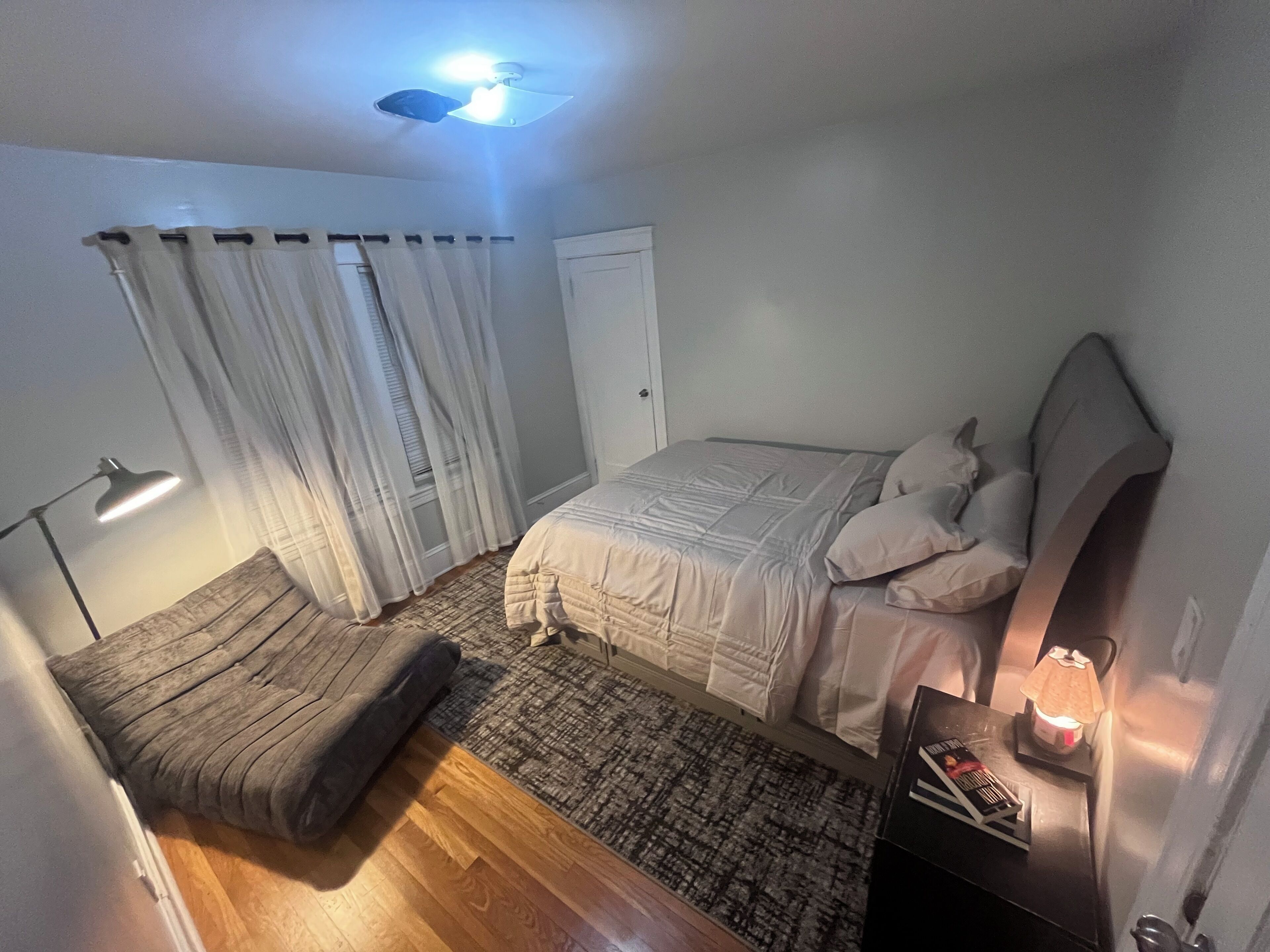 Un dormitorio con cama, mesita de noche, lámpara, ventana con cortinas, ventilador de techo y una alfombra pequeña.