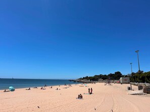 A wide sandy beach with people sunbathing and swimming, a clear blue sky, and a distant view of the sea. - Arcos House by the Sea, 1 bedroom Apt Sleeps 4 (Paço de Arcos)