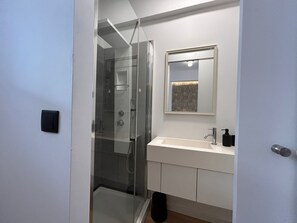 Une salle de bain moderne avec une douche à l’italienne, un meuble-lavabo blanc avec un miroir et une porte donnant sur une autre pièce.