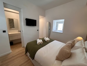 Ein Schlafzimmer mit Bett, Fernseher, Fenster, Spiegel und einem Badezimmer inklusive Waschbecken und Handtuchhalter.