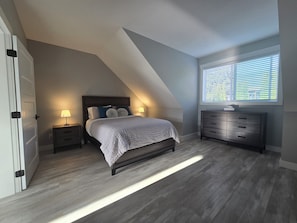 A bedroom with a sloped ceiling, a bed with a white bedspread, two bedside lamps, a dresser, and a window with blinds.