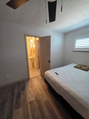Ein Schlafzimmer mit Bett, einem Fenster mit Jalousien, einer Tür zu einem Badezimmer und einem Deckenventilator.