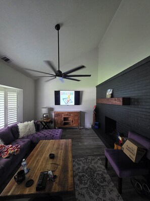 Ein Wohnzimmer mit Deckenventilator, Flachbildfernseher, Couchtisch aus Holz, violettem Sofa und Kamin.
