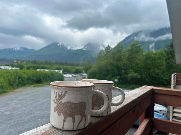 Deux tasses en céramique ornées de motifs animaliers, posées sur une rambarde en bois, avec vue sur les montagnes et la verdure.