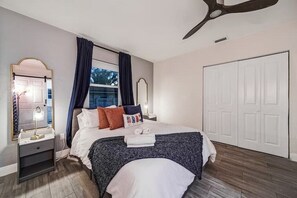 Ein Schlafzimmer mit Bett, Nachttisch, Spiegel, Fenster mit Vorhängen und einem Kleiderschrank.