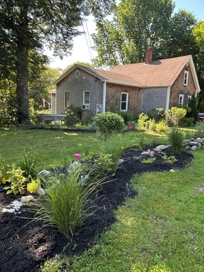 A two-story house with a brick exterior, a front yard with a variety of plants and a small garden bed, and a large tree to the left.