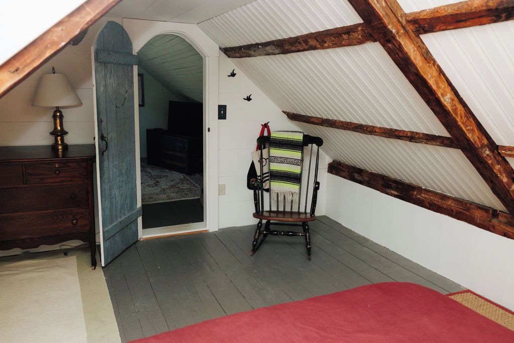 Une pièce avec un plafond en pente, une chaise en bois, une commode et une porte donnant sur une autre pièce.