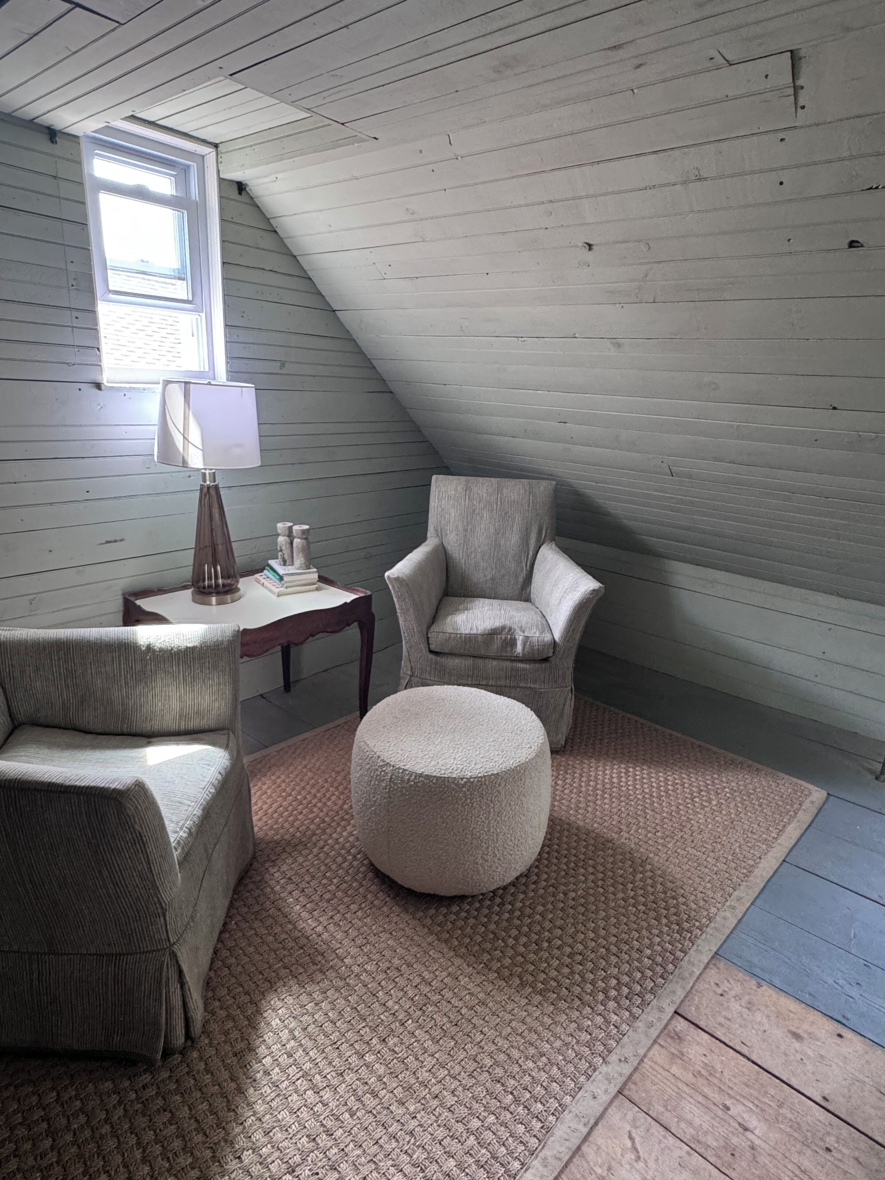 Une pièce de style loft avec un plafond en pente, une fenêtre, une lampe, une chaise, un canapé et un pouf rond.