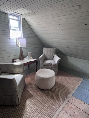 A loft-style room with a sloped ceiling, a window, a lamp, a chair, a sofa, and a round ottoman.