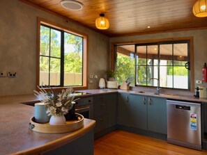 A modern kitchen with wooden cabinets, a stainless steel dishwasher, and large windows offering a view of the outdoors. - Stillwater Resort - Lakefront Estate, Sleeps 16 (Rotorua)