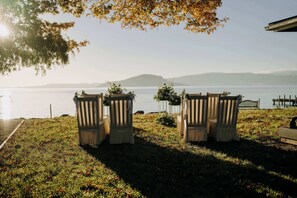 A lakeside setting with two wooden chairs and a bench, surrounded by grass and flowers.