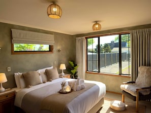A well-lit bedroom with a large bed, bedside lamps, a view of the outdoors through a window, and a chair with a fur throw. - Stillwater Resort - Lakefront Estate, Sleeps 16 (Rotorua)