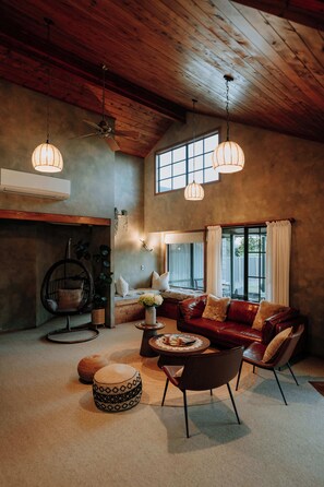 A spacious living room with a high ceiling, a leather sofa, a hanging chair, and a large window providing natural light. - Stillwater Resort - Lakefront Estate with two dwellings, spa pools, SUPs - Sleeps 16 (Rotorua)