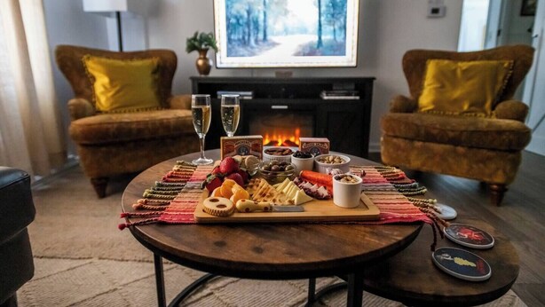 Uma sala de estar aconchegante com lareira, dois poltronas, uma mesa de centro redonda com petiscos e bebidas, e uma televisão exibindo uma cena de floresta.