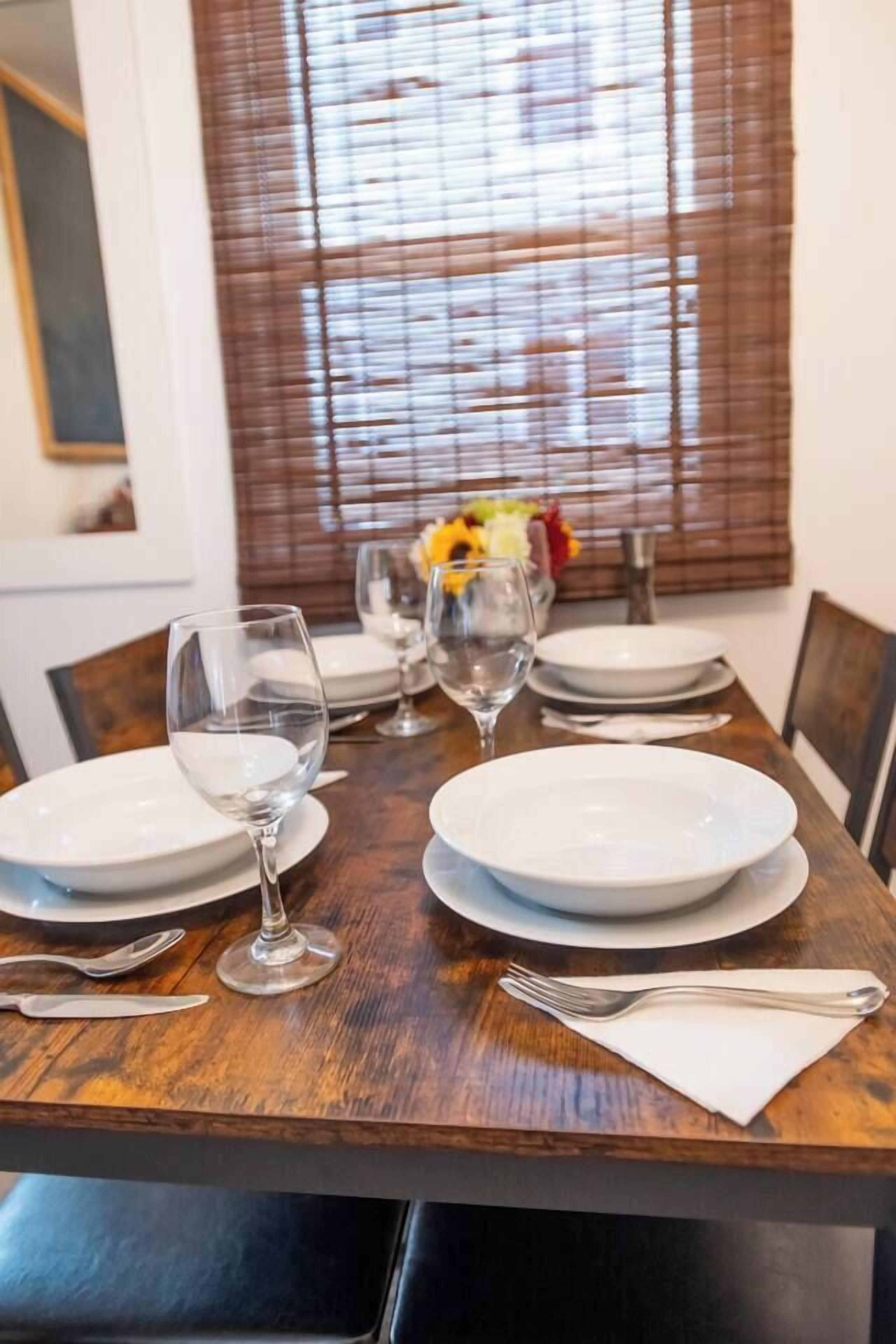 Una mesa de comedor dispuesta con platos, copas de vino y cubiertos, con una ventana y persianas al fondo.