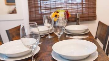 Una mesa de comedor dispuesta con platos, copas de vino y cubiertos, con una ventana y persianas al fondo.