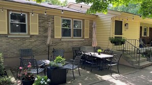 Um pátio com móveis de exterior, plantas em vasos e um edifício amarelo com janelas e porta.