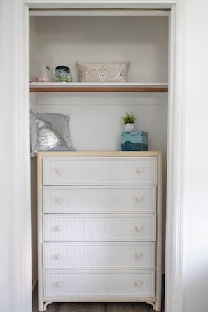 Ein weißer Korbuschrank mit Schubladen und Knöpfen, darüber ein Ablagefach mit Dekorationsgegenständen, und ein Wandregal mit einer Pflanze.