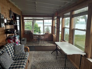 A cozy room with a wooden interior, a dining table with chairs, a sofa, and large windows offering a view of a lake.