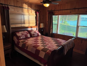 A wooden-paneled room with a bed, a ceiling fan, a window with blinds, and a view of greenery outside.