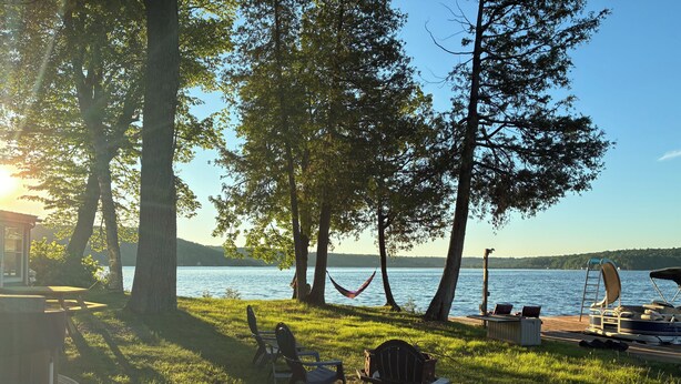 Ein Campingplatz am Seeufer mit einer Hängematte, Stühlen und einem Steg mit einem Boot.