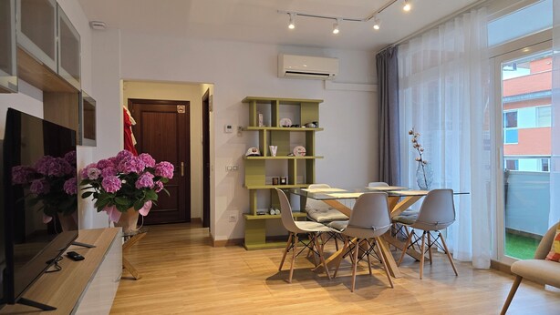 Una sala de estar moderna con televisor, piso de madera, zona de comedor con sillas, estantería verde y un ventanal con cortinas.