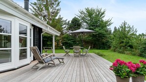 A wooden deck with outdoor furniture, a covered seating area, and a garden with trees and flowers. - 5 Star Holiday Home in Faxe Ladeplads (Faxe Ladeplads)