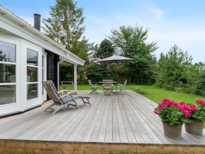 A wooden deck with outdoor furniture, a covered seating area, and a garden with trees and flowers. - 5 Star Holiday Home in Faxe Ladeplads (Faxe Ladeplads)