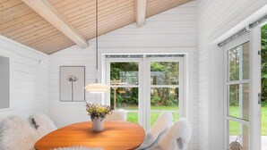 A dining area with a wooden table, white chairs, a vase of flowers, and a view of the outdoors through large windows. - 5 Star Holiday Home in Faxe Ladeplads (Faxe Ladeplads)