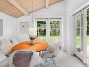 A dining area with a wooden table, white chairs, a vase of flowers, and a view of the outdoors through large windows. - 5 Star Holiday Home in Faxe Ladeplads (Faxe Ladeplads)