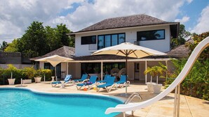 Une piscine avec des chaises longues et des parasols, une maison à deux étages dotée d’un balcon, et un ciel dégagé.