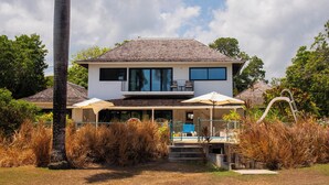 Une maison à deux étages avec un balcon, une terrasse couverte aménagée avec du mobilier d’extérieur et un grand arbre au premier plan.